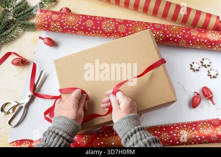 Mani che preparano un regalo, carta da imballaggio a motivi geometrici, nastro regalo, forbici e decorazioni natalizie su un tavolo, vista dall'alto, copia dello spazio sul Foto Stock