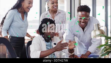 Tifo per un collega seduto che si dirige verso il laptop mentre il team applaude in ufficio, in camicie e cuffie Foto Stock