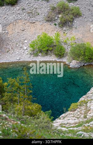 Colpo ad angolo alto guardando verso il basso in un fiume o in una piscina turchese cristallina che scorre accanto a una riva di ghiaia, con cespugli verdi e alberi sparsi. Foto Stock