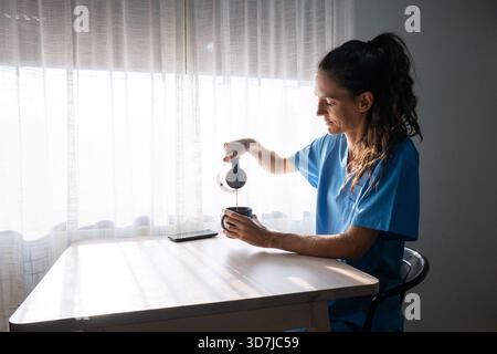 Donna in scrub che versa caffè in una tazza, facendo una pausa Foto Stock