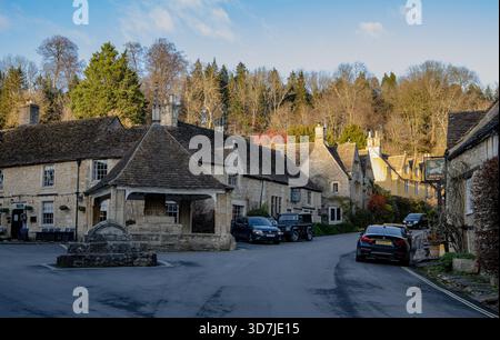 L'affascinante strada del villaggio a Castle Combe con la storica croce di burro di pietra e gli edifici, le automobili parcheggiate lungo la strada e il bosco sullo sfondo Foto Stock