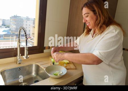 Donna in piedi in una cucina moderna, preparando felicemente verdure fresche su un tagliere accanto al lavandino, concentrandosi su cibo sano Foto Stock