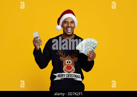 L'uomo con un maglione natalizio sorride mentre tiene in mano contanti e uno smartphone su sfondo giallo Foto Stock