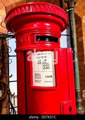 Scatola da posta a colonna in stile vittoriano inglese di colore rosso brillante con orari di raccolta esposti, su un'architettura in mattoni e barre in ferro per finestre Foto Stock