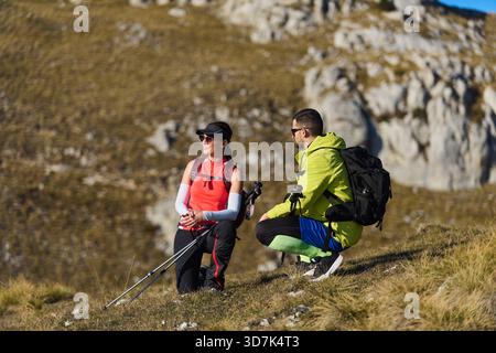 Due escursionisti si godono un momento di riposo su un soleggiato sentiero di montagna circondato da terreni rocciosi e vegetazione nel pomeriggio Foto Stock