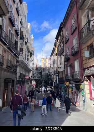 Vivace strada pedonale nel centro di Madrid con balconi storici, negozi e luci festose sotto un cielo blu brillante. Foto Stock