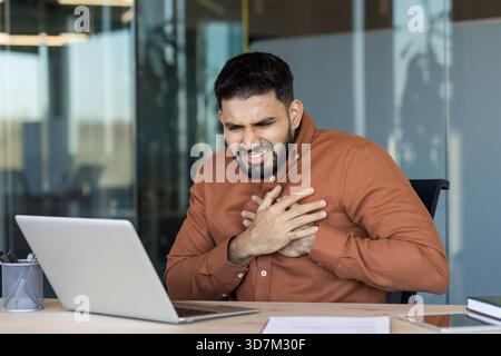 Uomo indiano che soffre di dolore toracico acuto mentre lavora su un portatile, indicando potenziali problemi cardiaci, stress o una crisi sanitaria in un ambiente aziendale Foto Stock