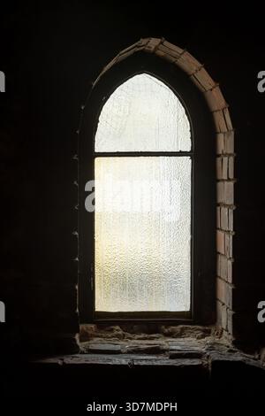Finestra ad arco in vetro smerigliato in una vecchia parete in mattoni con pannelli testurizzati, illuminata dalla luce del giorno dall'interno di una stanza scura con un arco gotico d'epoca Foto Stock