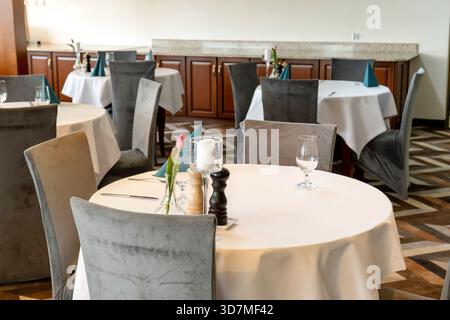 Tavoli rotondi con eleganti lenzuola bianche e sedie in velluto grigio in un'accogliente sala da pranzo del ristorante, ambienti con vetreria candele fiori pronti per l'uso Foto Stock
