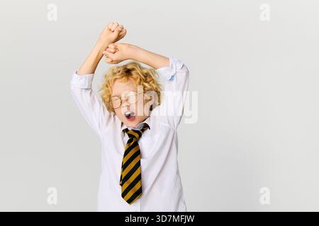 Un ragazzo felice con i capelli biondi ondulati si allunga indossando una camicia bianca e una cravatta a righe in uno studio. Foto Stock