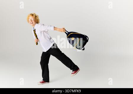 Un allegro ragazzo biondo in uniforme scolastica sorride mentre tiene in mano il suo zaino, pronto per la scuola. Foto Stock