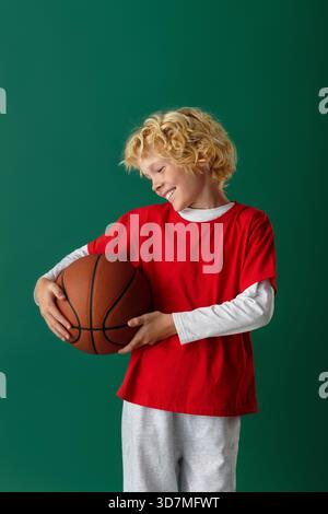 Un ragazzo sorridente in camicia rossa regge un pallacanestro mentre si trova su uno sfondo verde. Foto Stock