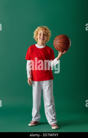 Un allegro biondo dai capelli ondulati si staglia su uno sfondo verde e regge una pallacanestro. Foto Stock