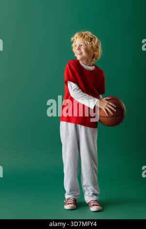 Un ragazzo biondo felice in abbigliamento casual rosso regala un pallacanestro su uno sfondo verde. Foto Stock