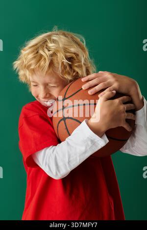 Un ragazzo biondo felice in abiti rossi sorride con una pallacanestro sullo sfondo verde. Foto Stock