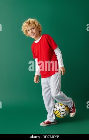 Un ragazzo felice con i capelli biondi ondulati indossa un abito rosso con una palla da calcio. Foto Stock