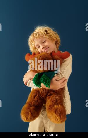 Un ragazzo biondo dai capelli ondulati tiene felicemente un peluche mentre indossa abiti casual. Foto Stock