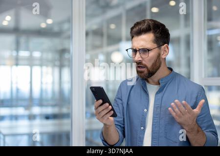 Uomo barbuto che indossa gli occhiali e una camicia blu che regge uno smartphone, esprimendo confusione e stress mentre guarda lo schermo in un moderno ambiente d'ufficio Foto Stock