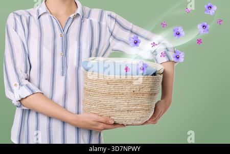 Donna che tiene in mano un cesto con abiti freschi e piacevolmente profumati su sfondo verde chiaro, primo piano. Flussi di aroma e fiori dal bucato. Utilizzo di fabr Foto Stock