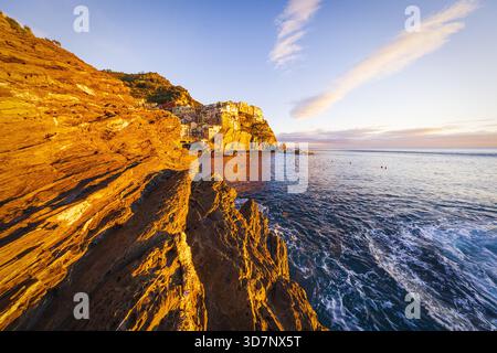 La vista della luce del sole dorata bagna le aspre scogliere e l'affascinante villaggio annidato sopra le onde turchesi che si infrangono, un paradiso costiero, Manarola, Liguri Foto Stock