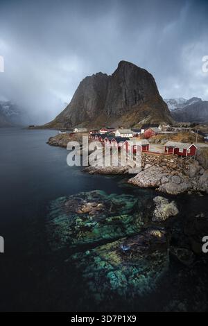 Vista delle cabine rosse annidate da acque turchesi sotto una montagna incombente in una giornata nuvolosa, l'essenza del fascino nordico, Reine, Nordland, Norvegia. Foto Stock