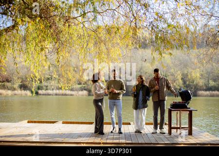 Quattro amici si riuniscono su un ponte di legno vicino al fiume, gustando bevande e cucinando su una griglia. La risata riempie l'aria sotto le foglie d'autunno dorate. Foto Stock