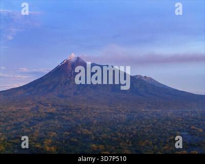 Veduta aerea del maestoso Monte Merapi che si erge sopra la lussureggiante foresta verde, la sua vetta avvolta da un delicato velo di nebbia, la Reggenza di Klaten, il centro di Giava, Foto Stock