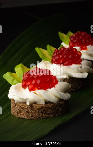 Tre torte rotonde condite con panna montata, caviale rosso brillante e fette di frutta verde sono disposte su una foglia di banana, creando un elegante piatto da dessert Foto Stock