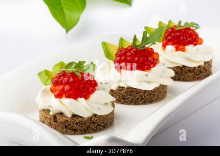 Tre splendidi antipasti sono disposti su un piatto bianco. Ognuna presenta una base di pane di segale ricoperto da ripieno cremoso, caviale rosso brillante e fresco Foto Stock