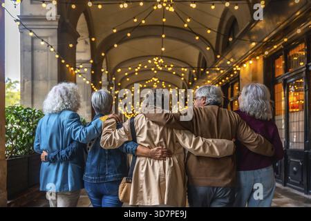 Un gruppo di diversi amici anziani che camminano insieme si abbracciano lungo un arco illuminato della città al crepuscolo, condividendo amicizia e godendosi un'escursione serale Foto Stock