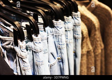 Primo piano di jeans e pantaloni in denim azzurro appesi a grucce nere accanto a un maglione in maglia beige in un negozio di abbigliamento. Foto di alta qualità Foto Stock