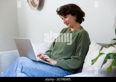 Donna seduta su un divano grigio chiaro che scrive su un computer portatile argento nel soggiorno con piante in vaso. Accogliente, moderno, minimalista, stile di vita, interni, spazio di lavoro, Foto Stock