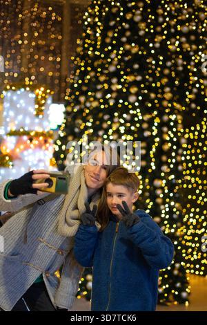 Madre e figlio fanno segnali di pace mentre fanno un selfie davanti a un albero di natale decorato Foto Stock