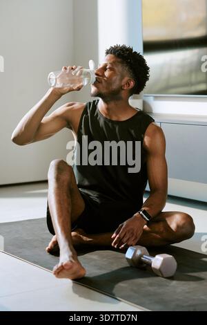 Giovane uomo Nero adulto seduto su tappetino da yoga con acqua potabile dopo l'allenamento mattutino Foto Stock
