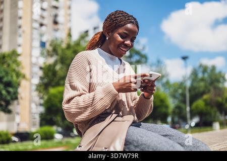 Donna afro-americana sorridente mentre invia SMS al telefono, crogiolandosi al sole, simboleggiando il tempo libero e la comunicazione. Foto Stock