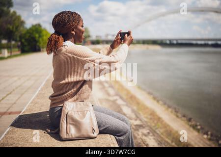 Giovane donna nera allegra sta scattando foto con lo smartphone. È seduta vicino al fiume in città. Foto Stock