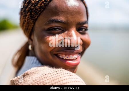 Primo piano ritratto di una giovane donna nera allegra che si gode una giornata di sole in città. Sta guardando la telecamera. Foto Stock