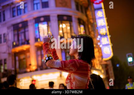 La donna fotografa di notte una strada luminosa della città con insegne al neon e un edificio storico. Shanghai, Cina Foto Stock