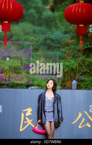 Donna in giacca di pelle che regge la borsa rossa si staglia accanto al muro con caratteri cinesi e lanterne rosse. Shanghai, Cina Foto Stock