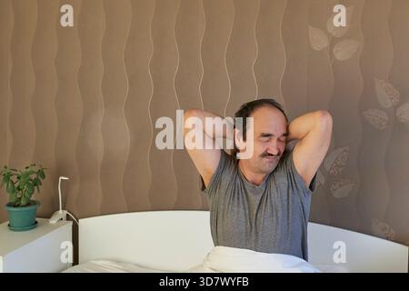 Un uomo maturo con i baffi, indossa una maglietta grigia, si siede a letto e allunga le braccia sopra la testa dopo una buona notte di sonno. La sua postura rilassata e il suo comfort Foto Stock