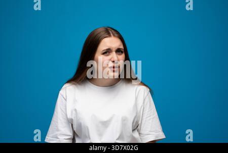Giovane donna con espressioni facciali confuse e scettiche in posa su sfondo blu in studio. Foto Stock