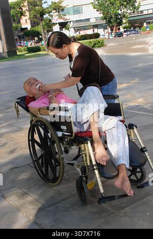 Una figlia amorevole o un caregiver si prende cura di un paziente anziano e gravemente malato in una sedia a rotelle all'aperto in un parco o in una piazza asiatica Foto Stock