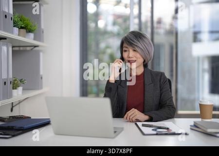 Donna professionista asiatica esperta impegnata attivamente in una conversazione telefonica mentre utilizza un computer portatile presso una scrivania moderna e luminosa. Foto Stock