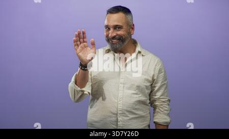Uomo ispanico di mezza età con barba al sale e al pepe sorridente in uno studio viola con camicia in lino beige e maniche arrotolate; amichevole fiducia. Foto Stock