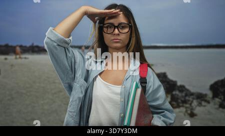 Donna che tiene in mano libri e zaino che protegge gli occhi con la mano in spiaggia indossando occhiali e camicia in denim mentre stringe quaderni e legante; studio giovanile d Foto Stock