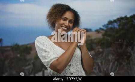 Donna che sorride e forma il cuore con le mani su una scogliera rocciosa che si affaccia sull'oceano; legame amoroso. Foto Stock