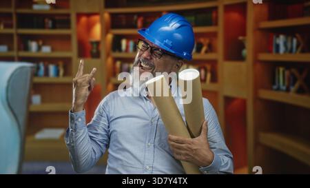 Uomo che indossa un elmetto blu che tiene in mano i piani arrotolati facendo gesti di corna di roccia nella costruzione; gioia celebrazione. Foto Stock