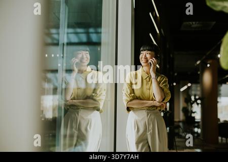 Una donna con una camicia gialla parla al telefono mentre si trova accanto a una grande finestra di vetro. La luce del sole mette in risalto il suo sorriso, creando un'atmosfera calda Foto Stock