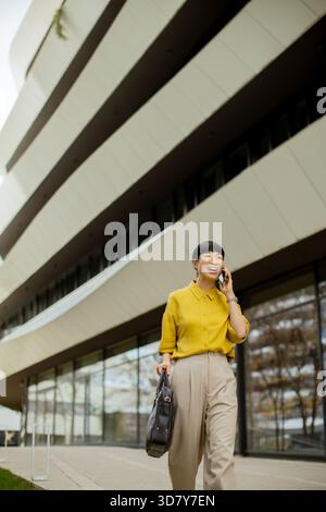 Una donna che indossa una camicetta gialla e pantaloni beige cammina felicemente fuori da un edificio moderno. Sta parlando al telefono, si sta godendo il bel tempo Foto Stock