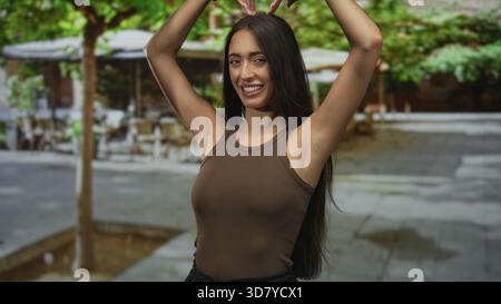 La giovane ispanica in canotta marrone forma il cuore con le mani e solleva le braccia sorridendo sulla terrazza di un ristorante di strada con alberi e tavoli; gioia Foto Stock